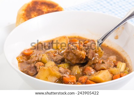 Spoon filled with large chunks of beef steak from bowl of beef stew.  Biscuit baked to golden brown and bright back light on blue gingham place mat.