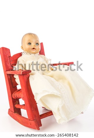 Antique baby doll sitting in old folk art  red rocking chair against white background; vertical format with copy space.