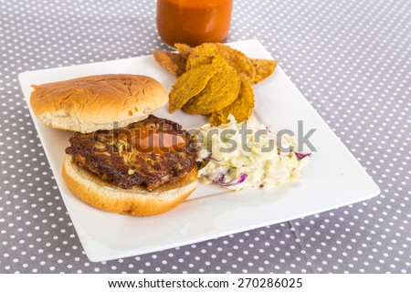 Crab Cake on toasted bun doused with hot sauce from bottle in background and served with sweet potato chips and coleslaw. Gray polka dot tablecloth with copy space.