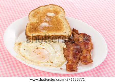 Egg over easy with fried bacon and buttered toast on pink gingham background.  Bright light from top right.