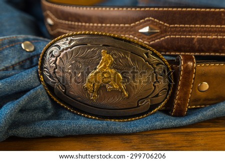 Closeup of Vintage silver buckle with cowboy on bucking bronc.  Leather belt with studs against blue denim button-snap work shirt background.