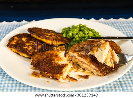 Chicken Cordon Bleu Dinner with potato pancakes and sweet peas.  Boneless chicken breast stuffed with ham and swiss cheese. White plate on blue gingham place mat against dark background.