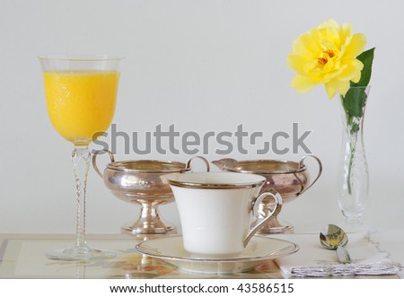 Place setting with orange juice in stemmed glass, coffee in fine china cup, and silver creamer and sugar bowl.  Isolated on pale gray and accented with a yellow rose.