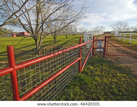 Beautiful evening light, White fences line this gravel road leading into a Texas Hill Country Ranch with bright red gate in foreground and red barn in the background