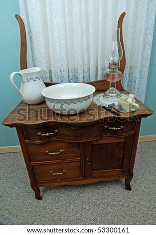 Old wash stand with with porcelain wash basin, pitcher, antique kerosene lamp, shaving mug and straight razor with white lace sheer curtains in background.