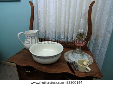 Beautiful porcelain pitcher and wash basin on antique wooden wash stand with kerosene lamp, shaving gear and sheer lace curtain in background