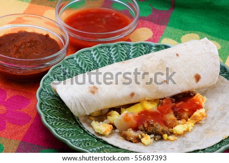 Egg, sausage and potato breakfast burritos on green plate setting on brightly colored place mat with individual servings of salsa and chili beans.