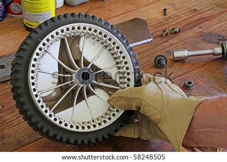 Tight shot of man's hand holding a damaged lawn mower wheel.  The plastic gear teeth are completely worn away.  Lawnmower blade, tools, nuts and bolts scattered on wooden workbench in background.