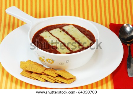 Chili bowl still bubbling hot with melted cheese and crackers on orange and yellow striped place mat with red napkin.