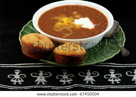 Bowl of spicy Tex-Mex chili and jalapeno cornbread with melted cheese and dollop of sour cream on black place mat with Aztec design.