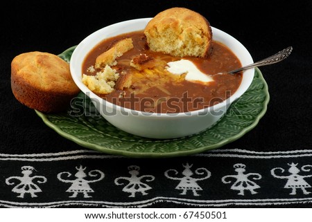 Bowl of spicy Tex-Mex Chili and jalapeno cornbread with melted cheese and dollop of sour cream against black Aztec design place mat.