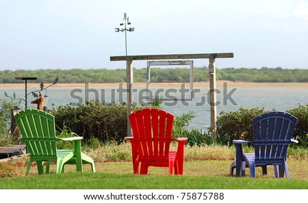 Relaxed setting with colorful lawn chairs overlooking a lake with garden and bird bath   Purple, green and red chairs.