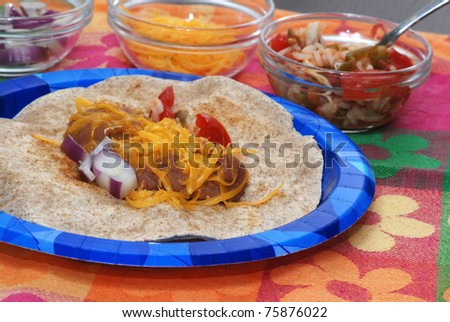 Bean and Cheese burrito on blue paper plate surrounded by Spanish Rice, cheese and red onion, sitting on colorful place mat.