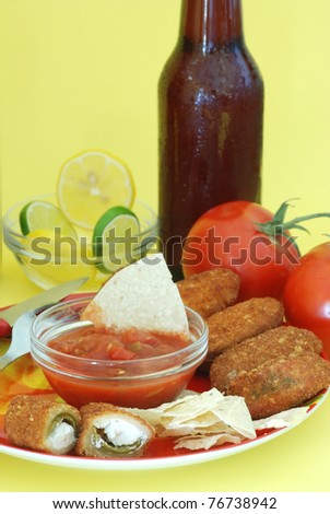 Jalapeno poppers on red plate with chips and salsa and surrounded by Mexican food ingredients; served with ice-cold beer on yellow background and party atmosphere.