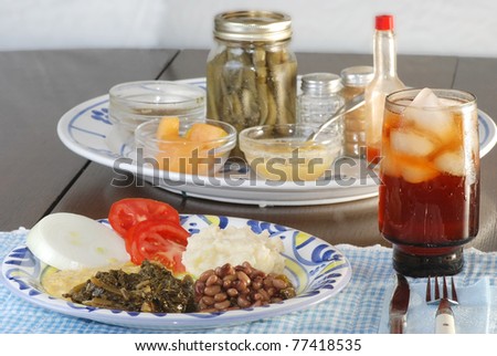 Place setting at dinner table with country style cooking; collard greens, black-eyed peas, creamed corn, mashed potatoes, sliced tomato and onion.  Hot sauce and pickled peppers on platter.