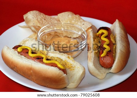 Two grilled hot dogs on white plate with chips and spicy cheese and jalapeno dip against red background.