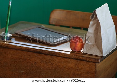 Old fashioned sack lunch and piece of fruit setting on antique school desk with vintage ink well, pencil, chalk board and book against green wall.