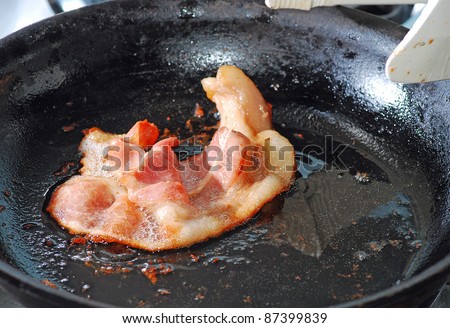 Single strip of Bacon sizzling in greasy skillet with white plastic tongs.