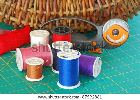 Wicker sewing basket is background for such needlecraft items as colorful spools of thread, pin cushion, rotary blade, cutting pad, and threaded needle.