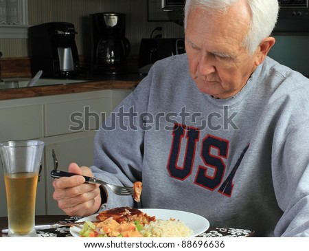 Old man at kitchen table alone dining on barbecue and beer.  Plate of pork tenderloin with bbq sauce, salad and rice against dark, depressing background.
