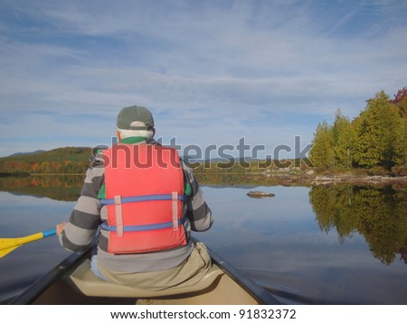Man with gray hair and bright orange life jacket paddling canoe on lake in Maine during colorful Fall Season.