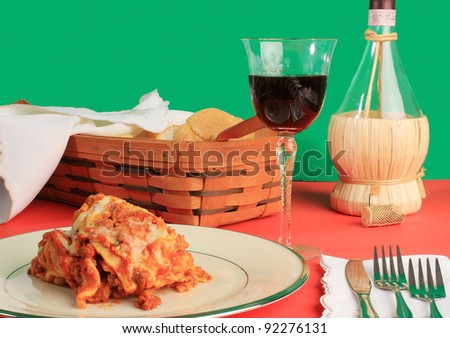 Lasagna on fine china with basket of garlic bread and bottle of chianti wine on red table with green background.