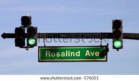 Two green traffic signals and a lighted street sign against a blue-sky background.