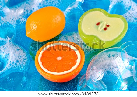 Three soaps in the shape of a lemon, orange, and apple in soap bubbles on a shiny watery blue background