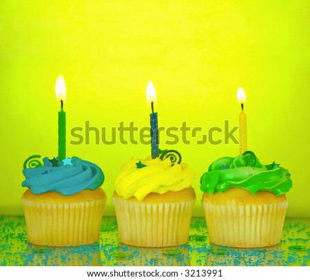 Three birthday cupcakes in blue, green, and yellow with lit candles, confetti, and sprinkles on a mirrored background