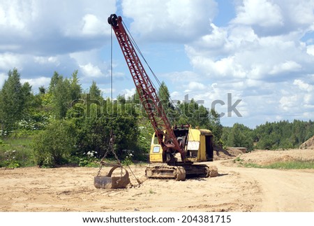 Yellow excavator with big heavy bucket standing on sand on background of forest and cloudy sky on summer day