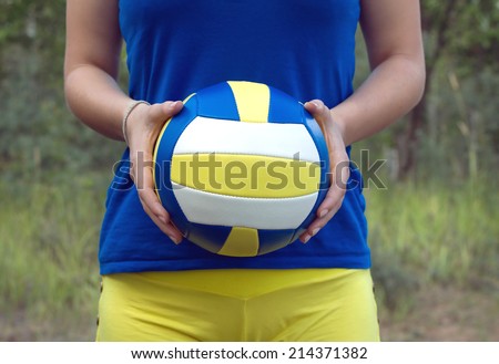 Teenage girl in blue shirt and yellow shorts holding a colorful sports ball for playing volleyball. Closeup Photo on blurred green background