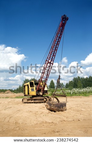 Yellow excavator with big heavy bucket standing on sand on background of forest and clear blue sky on a summer day vertical view