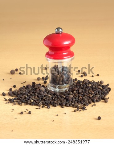 Still life with black peppercorn and manual mill with red cap, standing on brown wooden surface. Vertical photo closeup