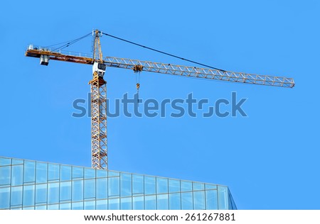 Yellow hoisting tower crane on top of construction skyscraper building over cloudless blue sky
