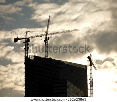 Contours of tower cranes above the highest office building under construction against cloudy sky sunset