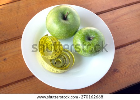 Yellow measuring tape and ripe green apples for weight diet lies on white plate on wooden table surface
