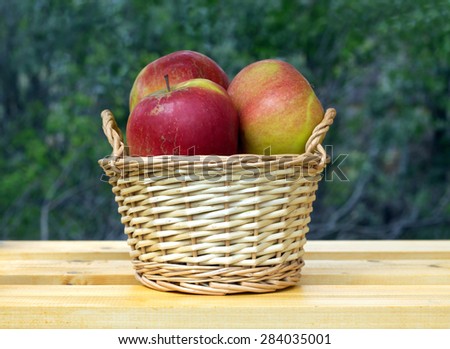 Ripe red and yellow apples in light brown wicker basket on wooden table against blur green background closeup
