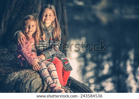 Little Sisters at the Lake. Small Caucasian Girls Hugging Each Other on the Large Old Tree Roots. Girls in the Park.