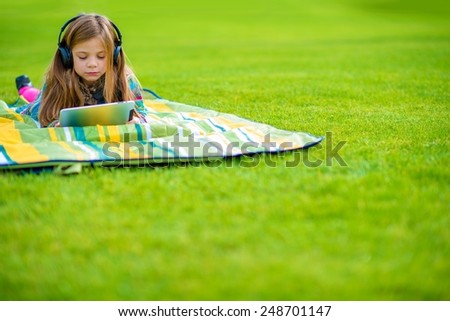 Girl Learning Language While Laying on the Park Green Field. Caucasian Girl with Wireless Headphones and Tablet.