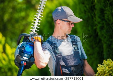 Garden Trimming Works. Professional Gardener in His 30s with Pro Hedge Trimmer Taking Care of the Garden.