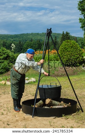 senior man preparing camping fire to cook corn in a big iron pot for corn party on the beach, Quebec, Canada