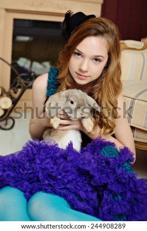 A young girl in the image of Alice in Wonderland is sitting near the fireplace and holds the rabbit
