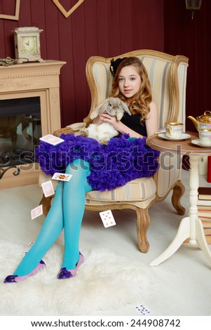 Young girl in the image of Alice in Wonderland is sitting in a chair near the fireplace and holds a rabbit