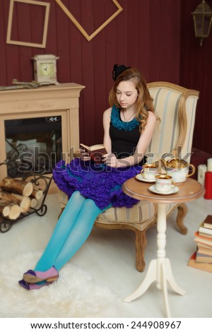 Young girl in the image of Alice in Wonderland is sitting in a chair near the fireplace and reading a book