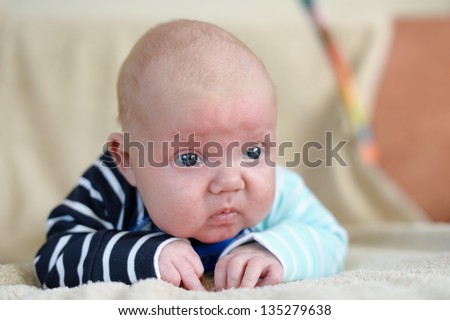 Cute 1,5 month old Caucasian baby boy with short blonde hair wearing a bright cloth and lying on his front on a bed