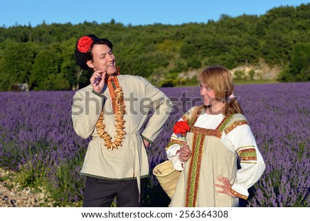Happy young couple having fun in a lavender field at sunset time. Warm light. Young man and beautiful girl in traditional russian clothes.