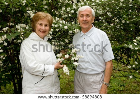 Elderly couple stands near a flowering bush in a garden. Smiles and looks in a photographic camera