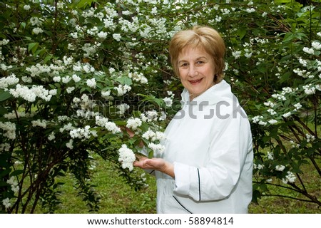 Elderly woman stands near a flowering bush in a garden. Smiles and looks in a photographic camera