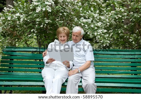 Elderly couple a  reads a  magazine.  Sits on a bench in a garden near a flowering bush