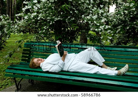 elderly woman reads a book lying on a bench in a garden near a flowering bush